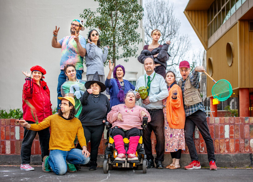 12 colourfully dressed people stand, sit and kneel around a low brick wall. Each have larger than life expressions on their faces. In the background are trees and buildings