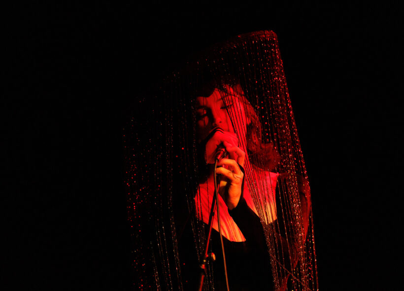 A singer lit in red and orange lighting holds a microphone close to their mouth. They are wearing a black long sleeve top and a large head piece with dangling strings of beads raining down their face. 
