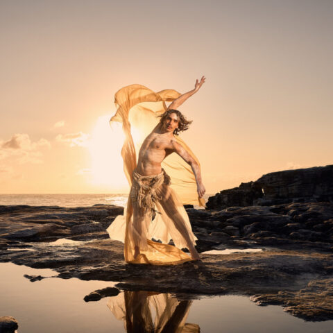Sunrise over the ocean. Clouds hover in the distance. Golden light flows across the sea and onto the rocky shoreline where a male dancer stands on the edge of some shallow rock pools.