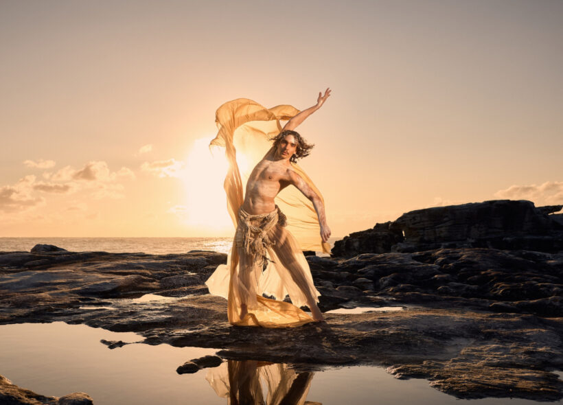 Sunrise over the ocean. Clouds hover in the distance. Golden light flows across the sea and onto the rocky shoreline where a male dancer stands on the edge of some shallow rock pools.
