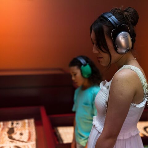 Two young people wear ear protection in a gallery space with muted lighting.