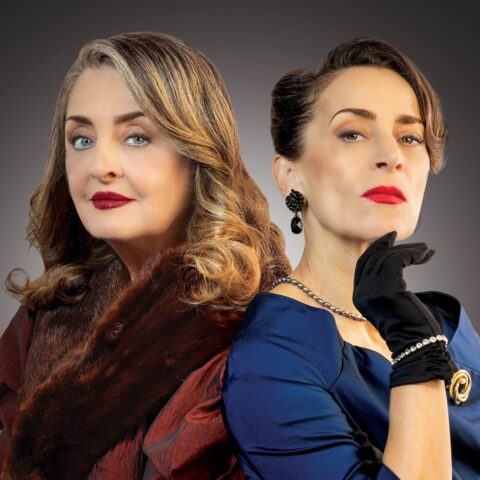 Two elegantly dressed women stand back-to-back against a dark gradient background. One wears a red dress with a fur stole, and the other a blue dress with gloves. Both have confident, poised expressions.