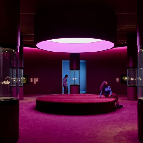 A photograph of one of the exhibition spaces. The room is a large circular space with deep pink and maroon furnishings. A large circular seat is in the center of the room, with cylindrical glass cases displaying tiaras. 