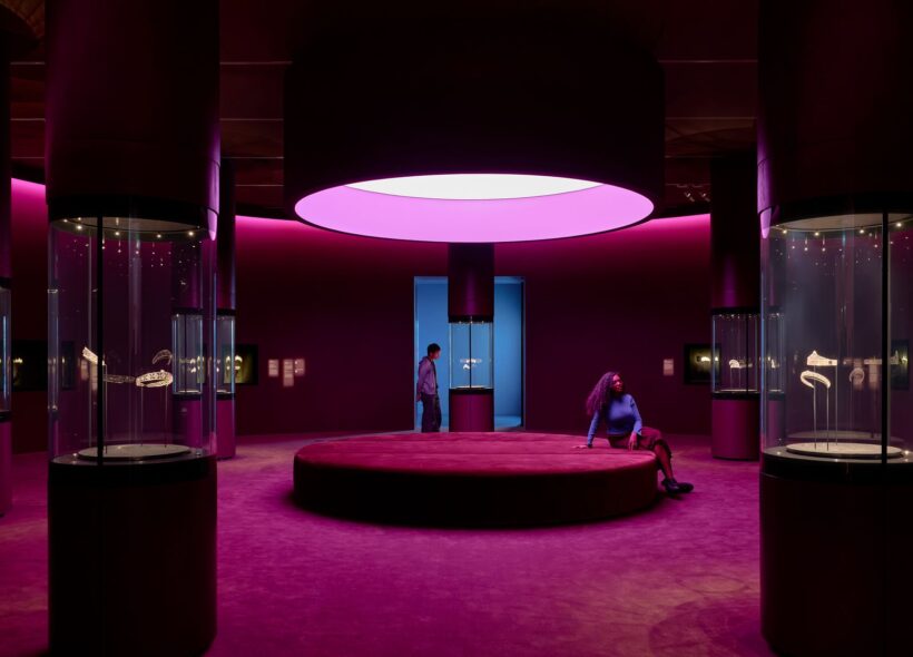 A photograph of one of the exhibition spaces. The room is a large circular space with deep pink and maroon furnishings. A large circular seat is in the center of the room, with cylindrical glass cases displaying tiaras. 
