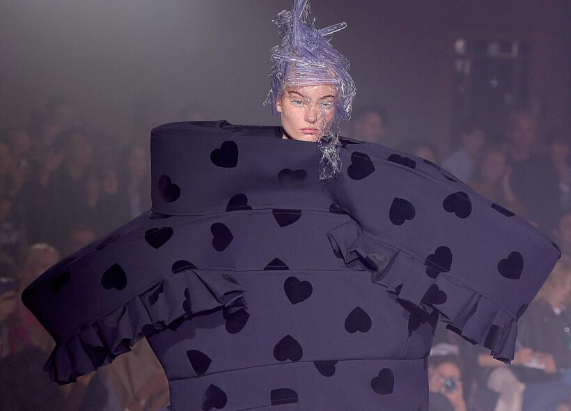 A photograph of a model on the runway wearing a Commes des Garcon design. The outfit is oversized on the model, with large bulky shapes protruding from their body. It is navy with darker blue or black love heart shapes across it and has some sections of frills. The model wears a plastic head piece that covers part of their face. 