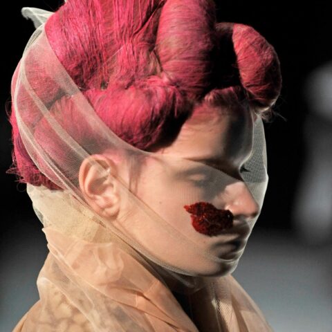 A close view of a model wearing a Commes des Garcon design. Their hair appears wrapped in bright red string and styled in voluminous shapes. Their face is partially covered by a sheer netting with a red lip shape embraided onto it.