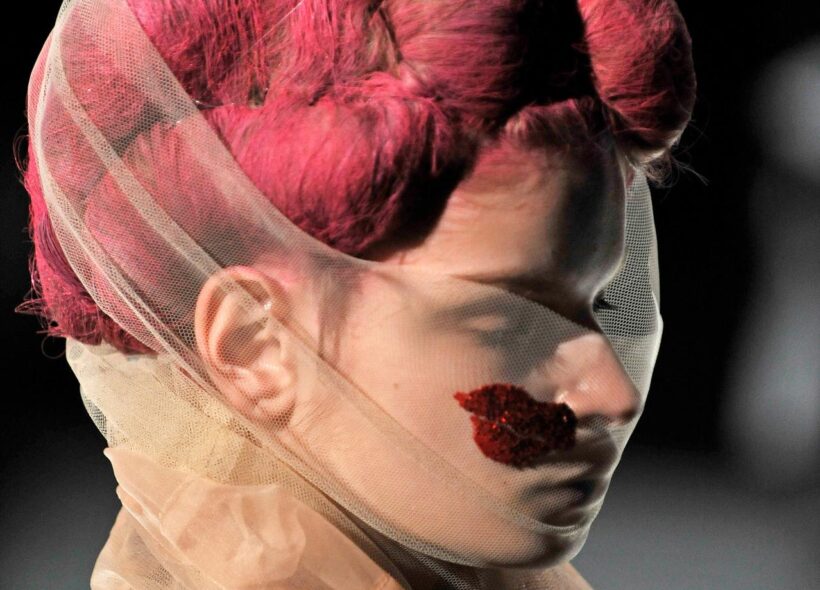 A close view of a model wearing a Commes des Garcon design. Their hair appears wrapped in bright red string and styled in voluminous shapes. Their face is partially covered by a sheer netting with a red lip shape embraided onto it.