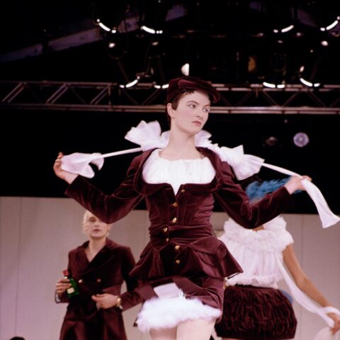 Models walk down the runway. In front, a pale-skinned model with cropped hair wears a deep maroon tailored dress with gold buttons. A white shirt and skirt peep out from underneath. The model is removing a white ruff or shawl from their shoulders as they walk. 