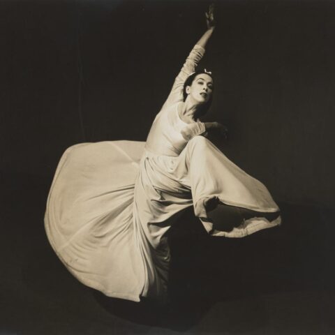 A black and white photograph of a woman dancing. She wears a long sleeved white or cream dress with a full skirt. The skirt flares around her as she moves. Her right foot is raised with her right elbow touching, her left arm is stretched up above. 