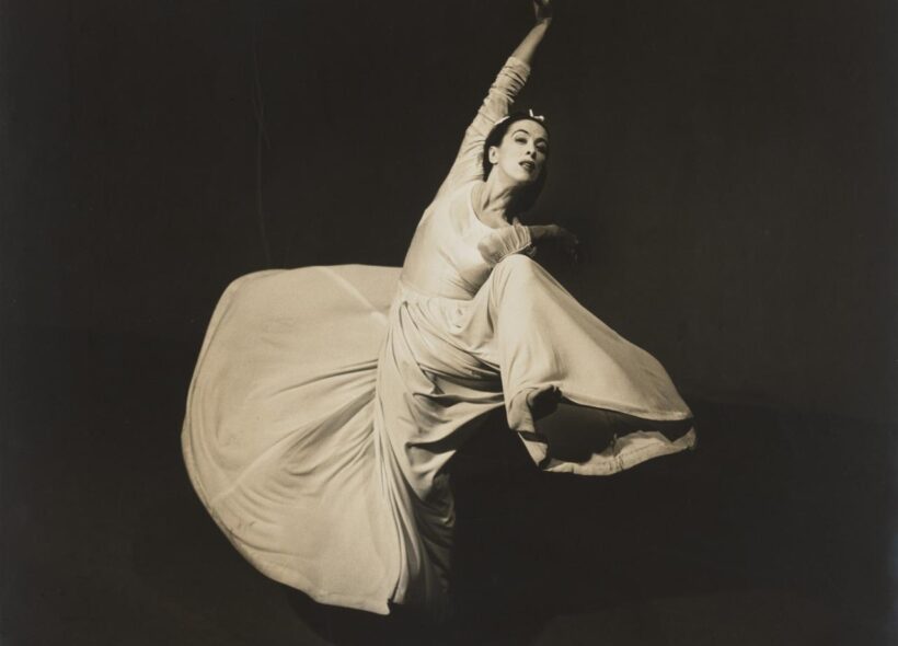 A black and white photograph of a woman dancing. She wears a long sleeved white or cream dress with a full skirt. The skirt flares around her as she moves. Her right foot is raised with her right elbow touching, her left arm is stretched up above. 