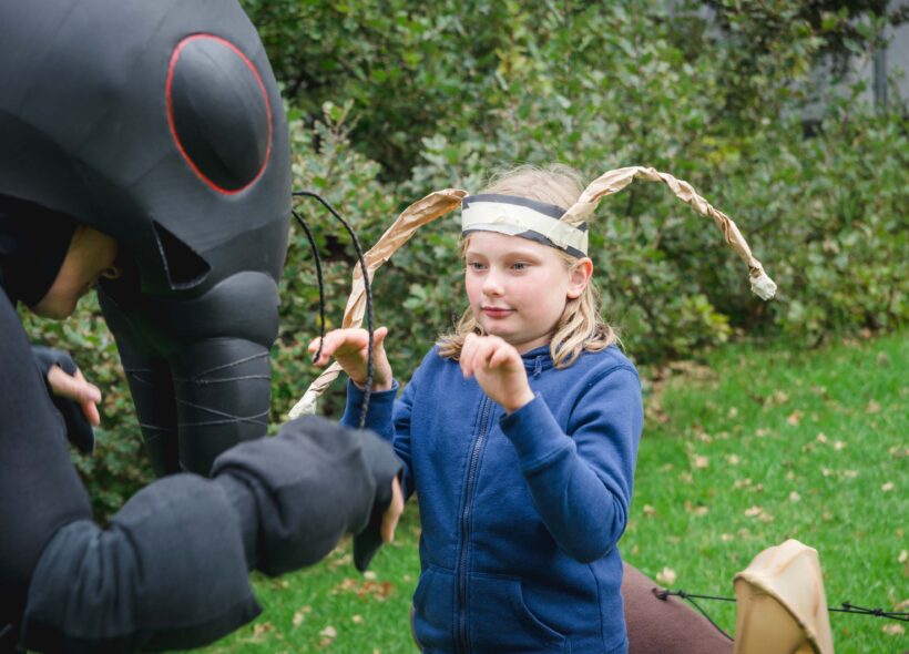 An outdoor Ants production photo. A Polyglot artist in an intricate Ant costume interacts with a child wearing a blue jumper and handmade paper antennae. Green grass and shrubs are visible in the background.