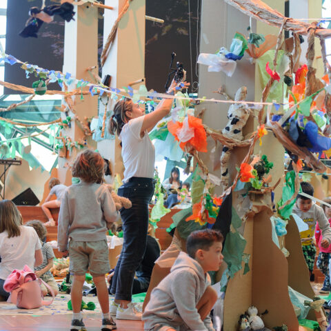 Paper Planet production photo: Sarah Walker. A forest of tall brown cardboard trees adorned with colourful tissue paper creatures and plants grows in a large space filled with natural light. Children and their families create and play with paper and tape among the trees.