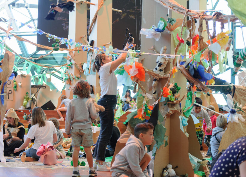 Paper Planet production photo: Sarah Walker. A forest of tall brown cardboard trees adorned with colourful tissue paper creatures and plants grows in a large space filled with natural light. Children and their families create and play with paper and tape among the trees.
