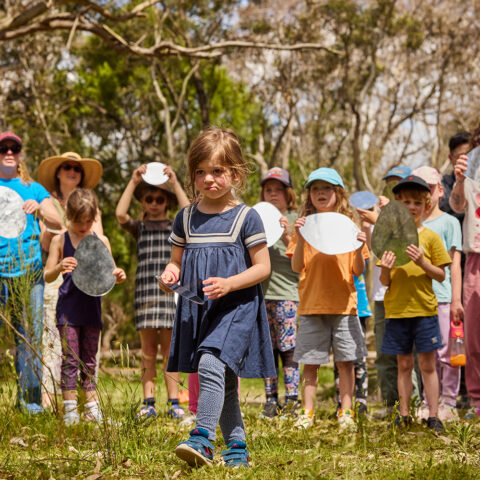 Forest workshop photo: Laura May Grogan. A child wearing a navy-blue dress is centred in the frame, walking into the forest. There are other children and adults standing in a line behind them, all holding circular mirrors. There are trees in the background.