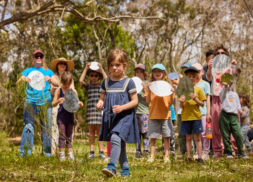 Forest workshop photo: Laura May Grogan. A child wearing a navy-blue dress is centred in the frame, walking into the forest. There are other children and adults standing in a line behind them, all holding circular mirrors. There are trees in the background.