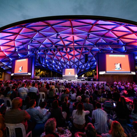  An audience gathers in an open-air amphitheater under a geometric dome lit with vibrant purple and red lights, attentively watching a large screen.