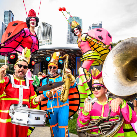 Colorful performers dressed as insects, playing instruments. Vibrant, joyful atmosphere with city skyline in the background.