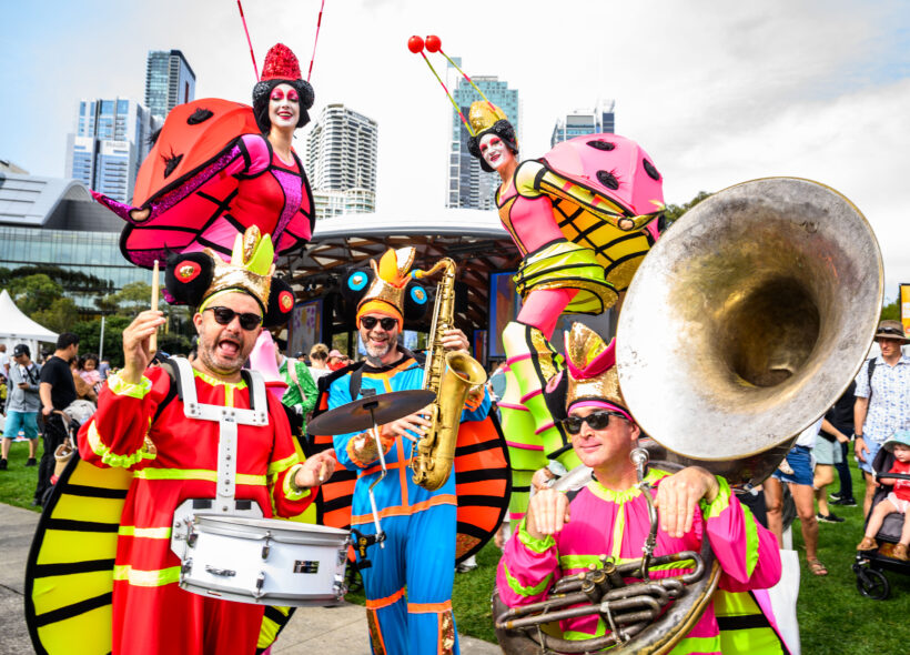 Colorful performers dressed as insects, playing instruments. Vibrant, joyful atmosphere with city skyline in the background.