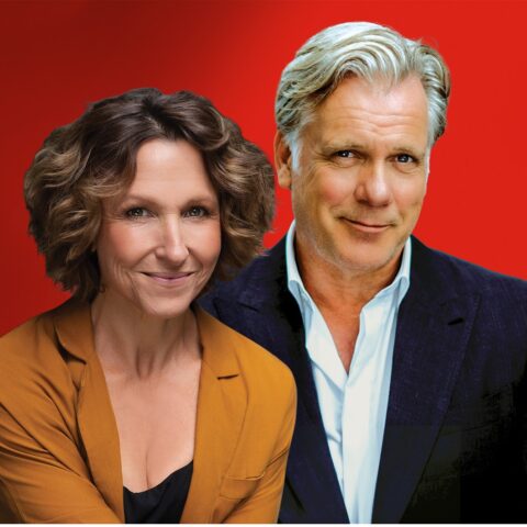 A woman in an orange blazer and a man in a navy suit pose together against a vibrant red background, both smiling warmly at the camera.