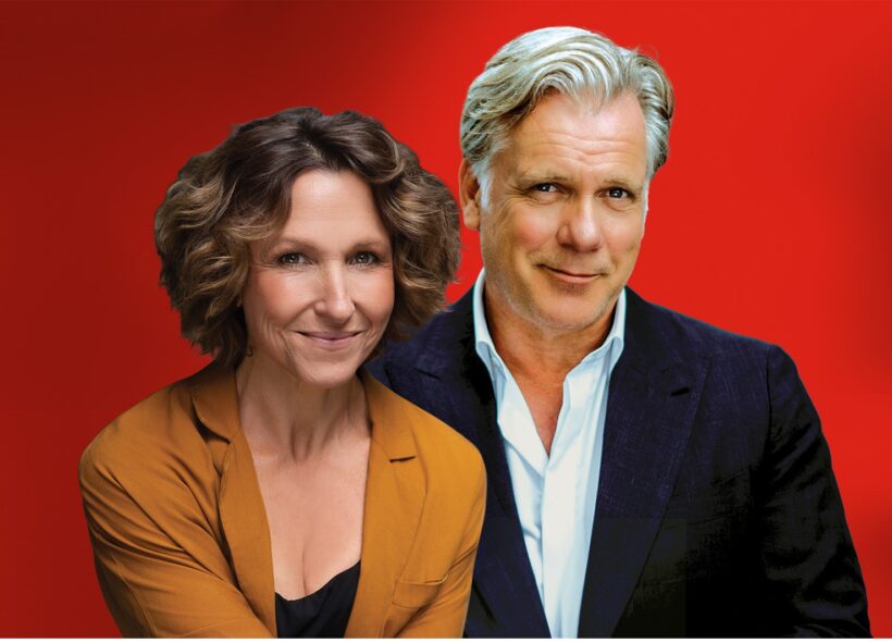 A woman in an orange blazer and a man in a navy suit pose together against a vibrant red background, both smiling warmly at the camera.