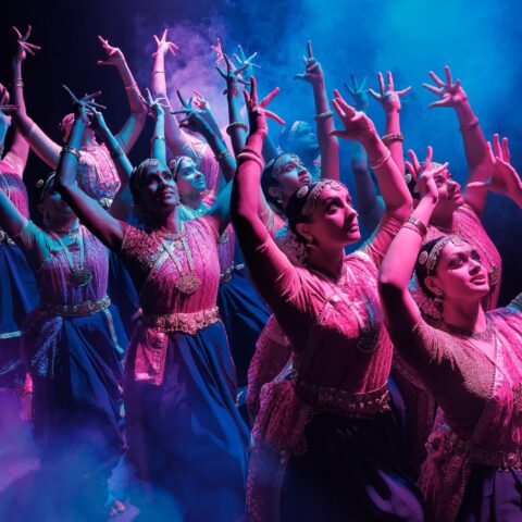 A group of dancers in traditional attire strike graceful poses, their hands elegantly lifted. Dramatic pink and blue lighting adds a mystical atmosphere.