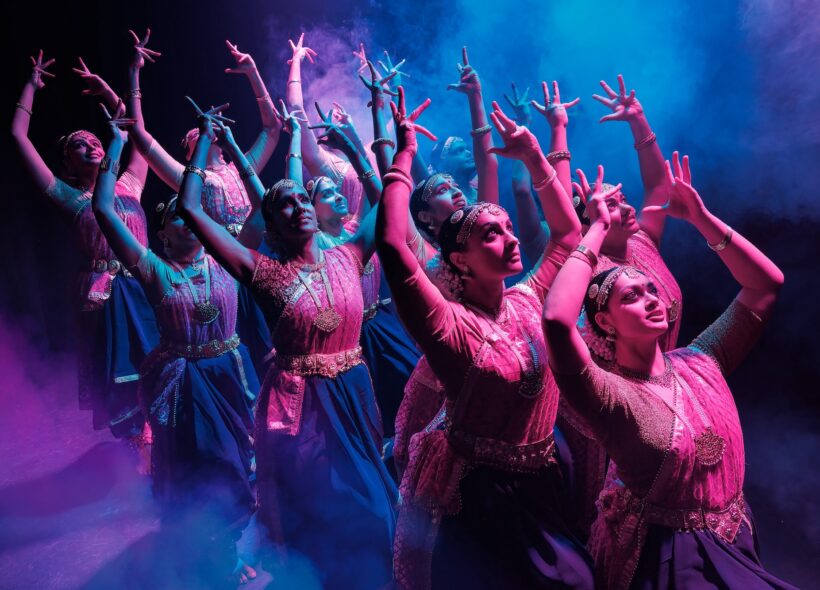 A group of dancers in traditional attire strike graceful poses, their hands elegantly lifted. Dramatic pink and blue lighting adds a mystical atmosphere.