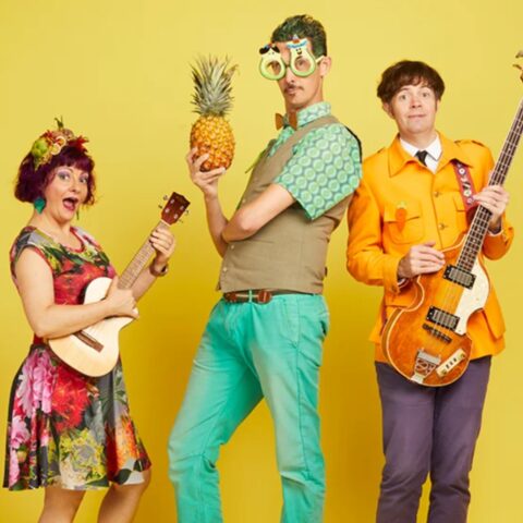 Three people pose against a yellow background; one with a ukulele in a floral dress, one holding a pineapple, wearing quirky glasses, and one in an orange jacket with a bass guitar. The scene is playful and colorful.