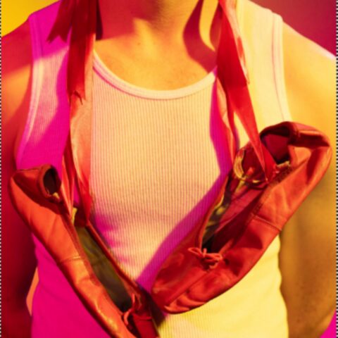 A person wearing a white tank top holds red ballet shoes slung around their neck. The vibrant pink and yellow lighting creates a warm, artistic tone.