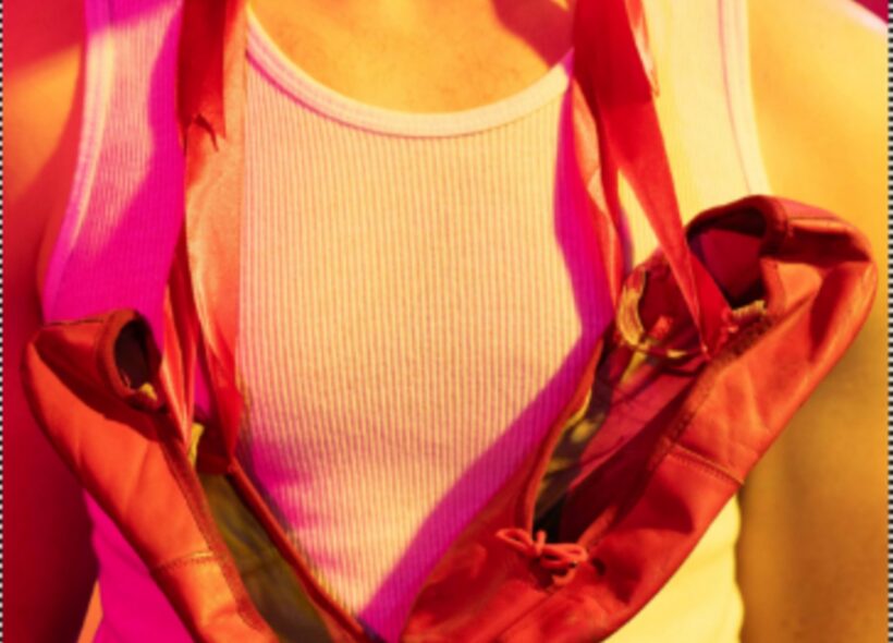 A person wearing a white tank top holds red ballet shoes slung around their neck. The vibrant pink and yellow lighting creates a warm, artistic tone.