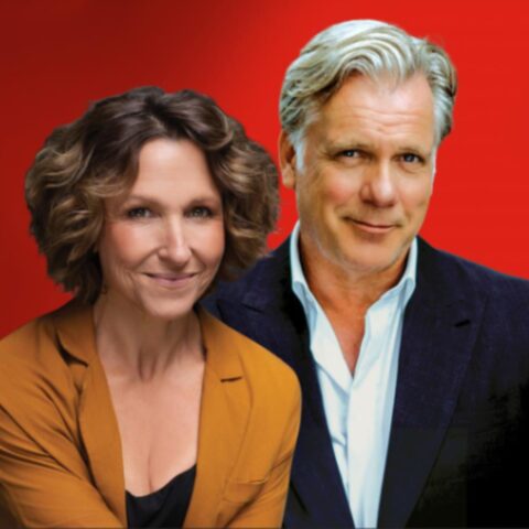 A woman in an orange blazer and a man in a navy suit pose together against a vibrant red background, both smiling warmly at the camera.