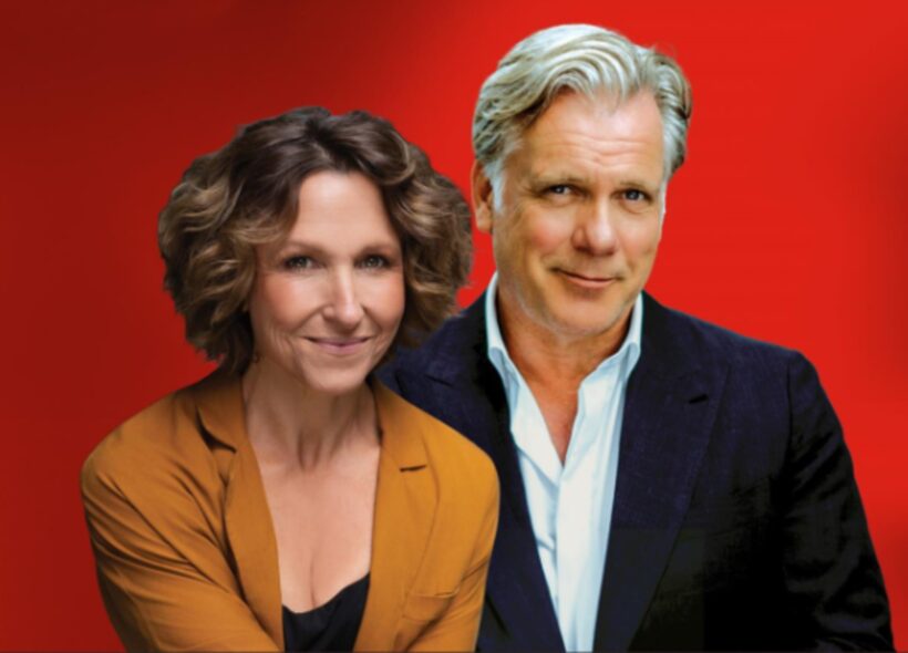 A woman in an orange blazer and a man in a navy suit pose together against a vibrant red background, both smiling warmly at the camera.