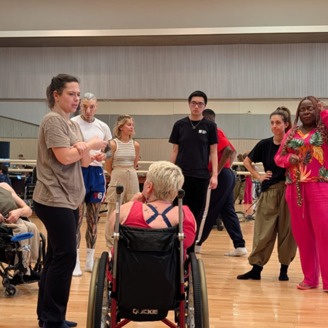 A group of dancers gathered in a studio, standing in a circle and listening to a woman speaking.
