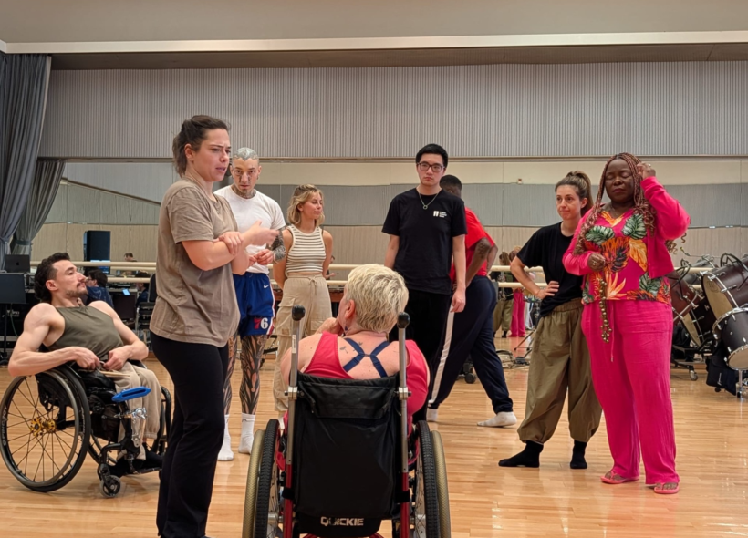A group of dancers gathered in a studio, standing in a circle and listening to a woman speaking.
