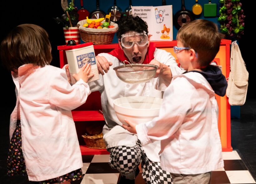 A person in goggles, holding a sieve over a mixing bowl, looks surprised in a playful kitchen scene with two children in white coats assisting.