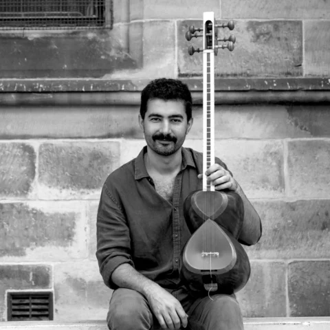 A man sits on stone steps, smiling and holding a traditional string instrument vertically. The background features a textured stone wall.