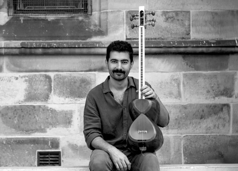 A man sits on stone steps, smiling and holding a traditional string instrument vertically. The background features a textured stone wall.