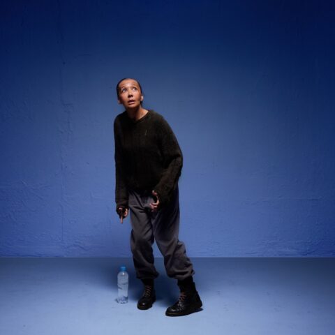 A person in casual attire stands in a blue-lit room, looking upward with a contemplative expression. A water bottle is on the floor beside them.