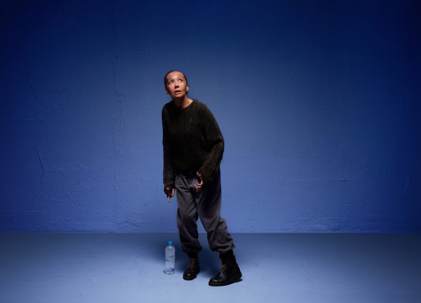 A person in casual attire stands in a blue-lit room, looking upward with a contemplative expression. A water bottle is on the floor beside them.