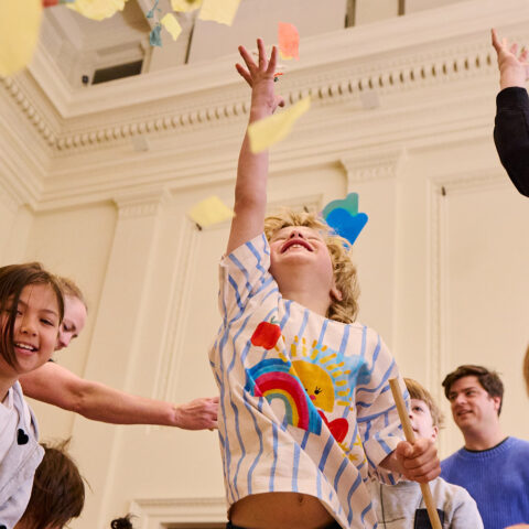 Whirlwind production photo: Laura May Grogan. A child jumps into the air, reaching for swirling colourful paper confetti. Other adults and children reach, jump and move around them.