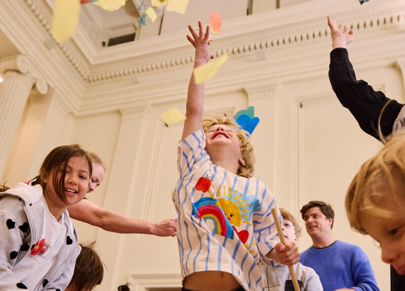 Whirlwind production photo: Laura May Grogan. A child jumps into the air, reaching for swirling colourful paper confetti. Other adults and children reach, jump and move around them.