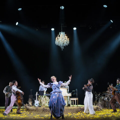 A theatrical performance featuring a joyful cast in vintage attire on a dimly lit stage. A chandelier and bright spotlights enhance the lively scene.