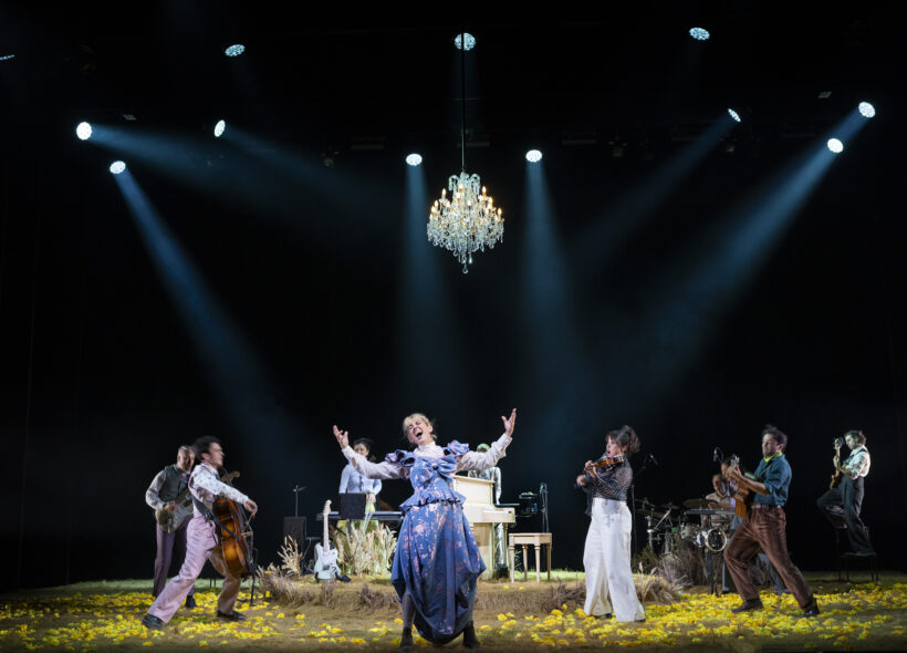A theatrical performance featuring a joyful cast in vintage attire on a dimly lit stage. A chandelier and bright spotlights enhance the lively scene.