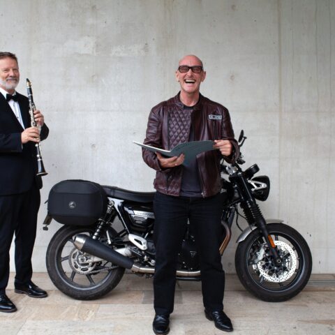 A smiling man in a leather jacket holds a folder in front of a motorcycle. A man in a tuxedo plays a clarinet beside him, against a concrete wall.