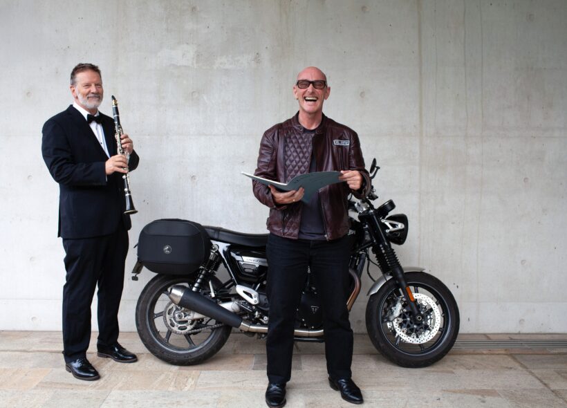 A smiling man in a leather jacket holds a folder in front of a motorcycle. A man in a tuxedo plays a clarinet beside him, against a concrete wall.