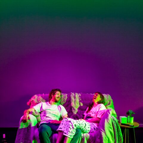 A couple laughs together on a sofa under vibrant green and purple lighting, creating a lively, joyful atmosphere. A wine bottle and shoes are on the floor. 