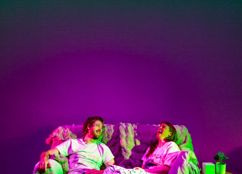 A couple laughs together on a sofa under vibrant green and purple lighting, creating a lively, joyful atmosphere. A wine bottle and shoes are on the floor. 