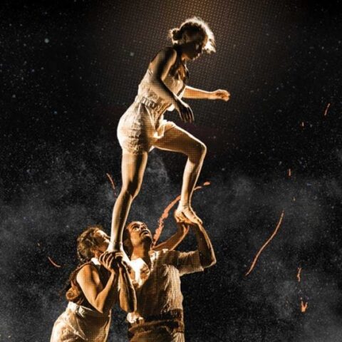   Three acrobats perform on stage under a spotlight, with two lifting a third person high. They wear light costumes, conveying elegance and balance.