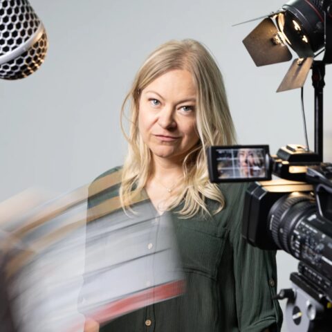 A woman with blonde hair stands in a studio setting, surrounded by filming equipment. She's in focus, conveying a professional and focused atmosphere.