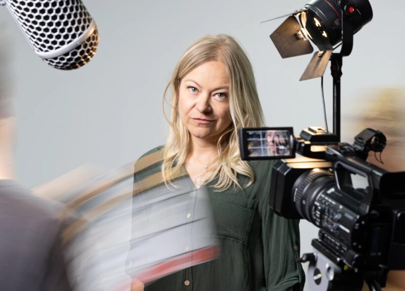 A woman with blonde hair stands in a studio setting, surrounded by filming equipment. She's in focus, conveying a professional and focused atmosphere.