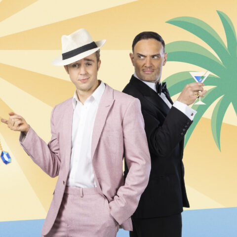 One man in a pink suit and fedora holding a diamond on a chain standing beside another man in a black tie suit holding a martini